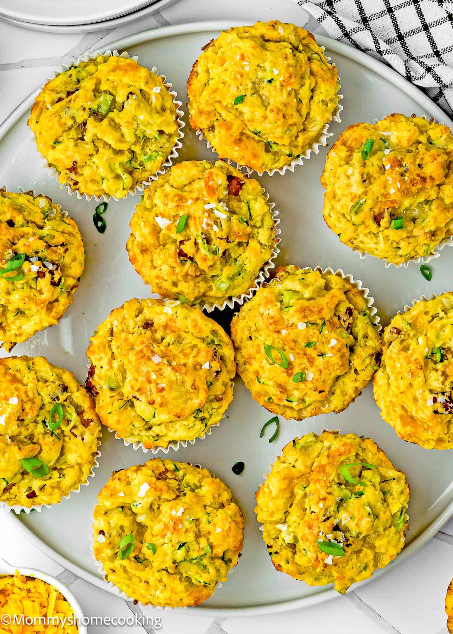 A plate of Eggless Cheesy Savory Muffins, topped with chopped green herbs and sea salt, arranged in white paper liners on a white plate.