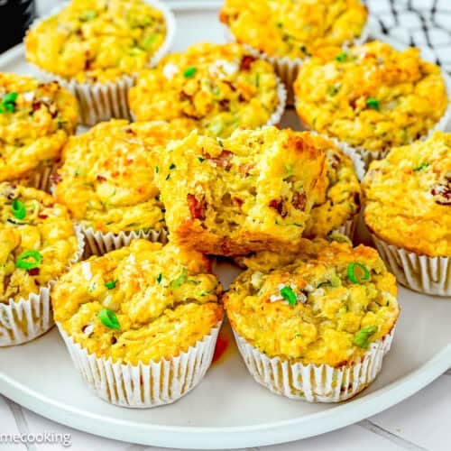 A plate of Eggless Cheesy Savory Muffins with a golden crust, garnished with chopped green herbs, displayed in white paper liners.