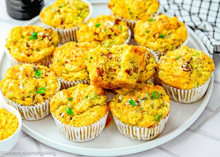 A plate of Eggless Cheesy Savory Muffins with a golden crust, garnished with chopped green herbs, displayed in white paper liners.