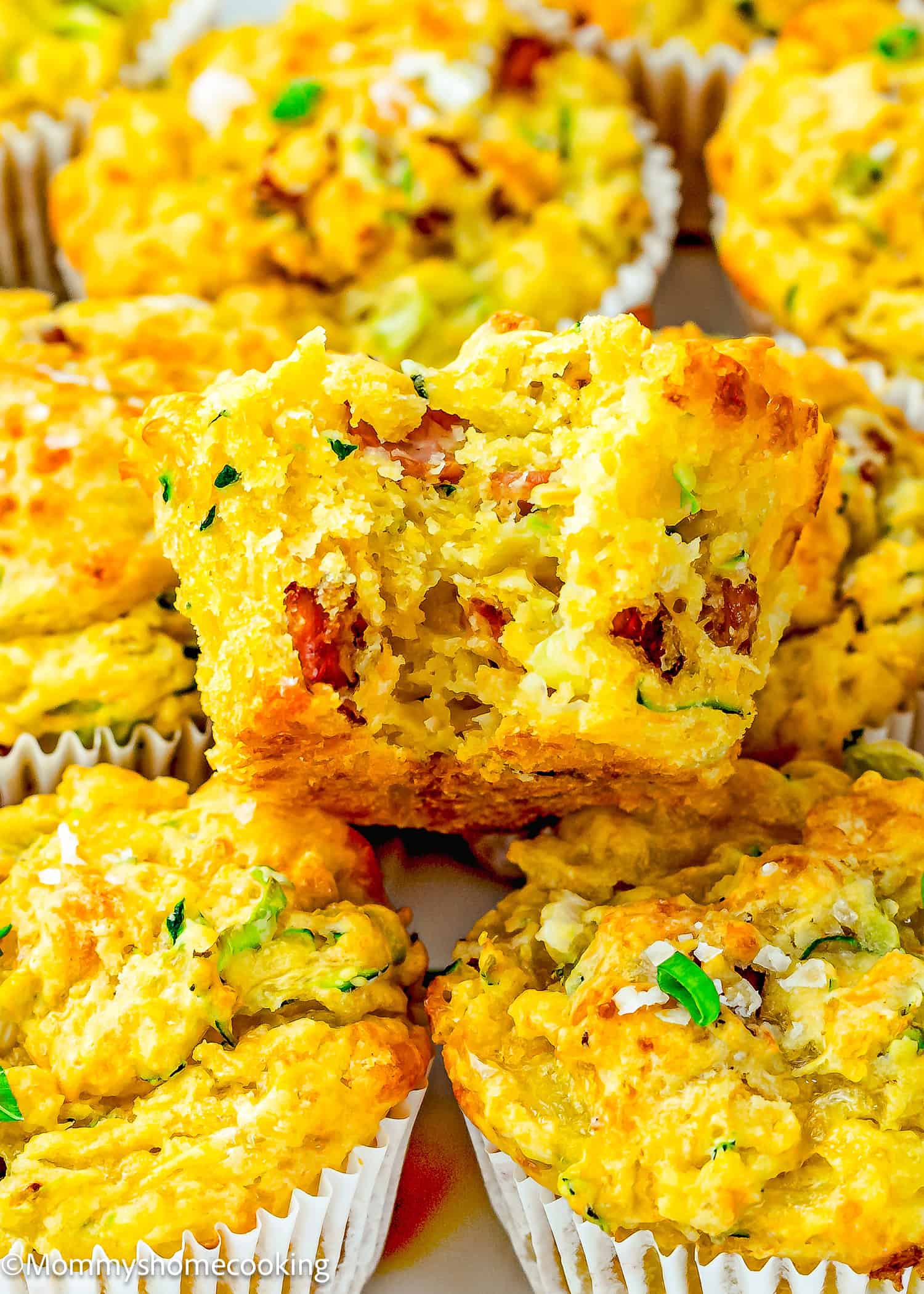 Close-up of Eggless Cheesy Savory Muffins with shredded zucchini and bacon pieces, one muffin broken in half to reveal the moist interior, all arranged in white paper liners.