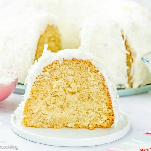 A slice of Eggless Coconut Bundt Cake with white frosting and shredded coconut sits on a plate, with the rest of the cake and ingredients visible in the background.