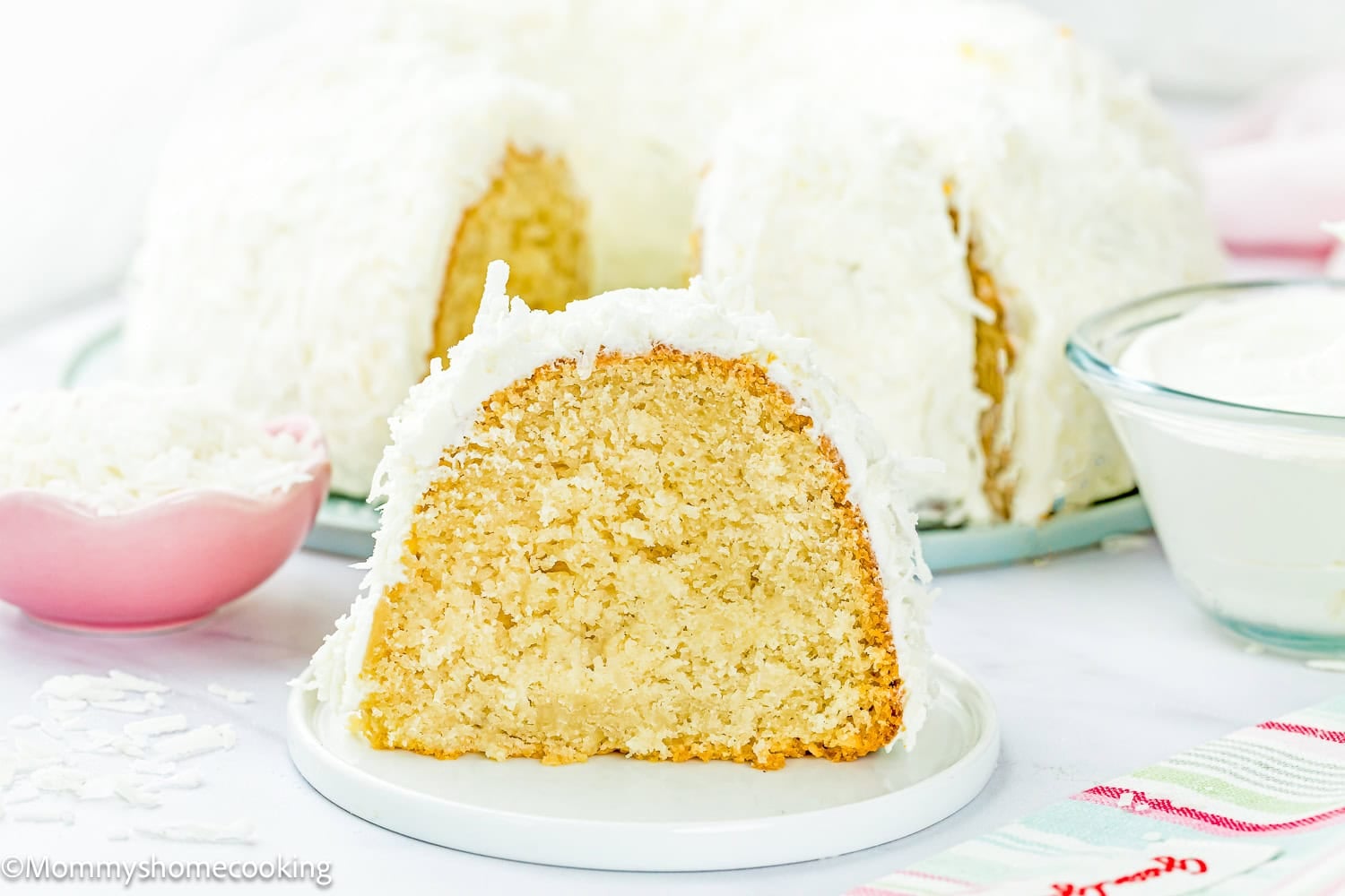 A slice of Eggless Coconut Bundt Cake with white frosting and shredded coconut sits on a plate, with the rest of the cake and ingredients visible in the background.