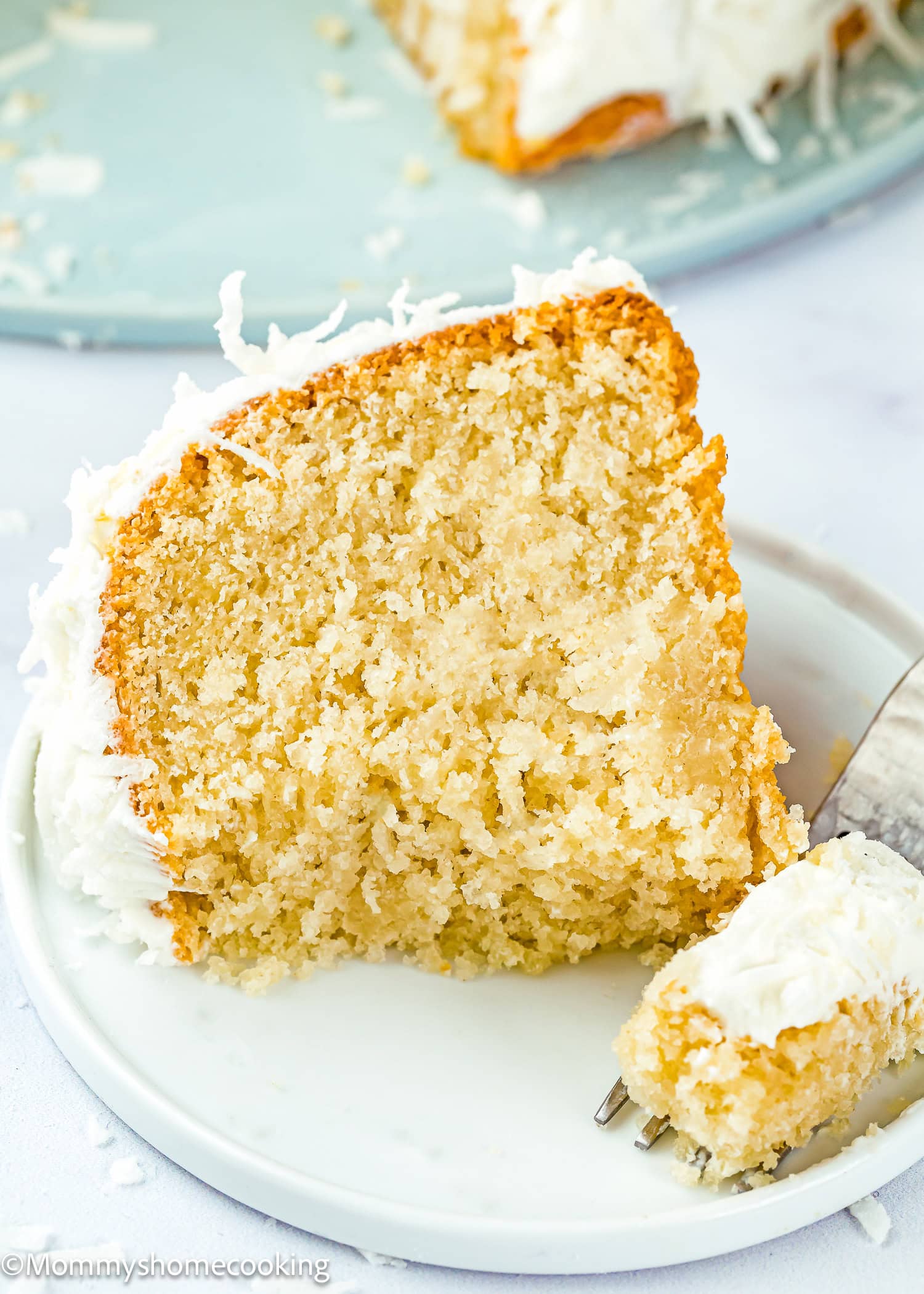 A slice of Eggless Coconut Bund Cake with white frosting sits on a plate, while a fork holds a small piece in front.
