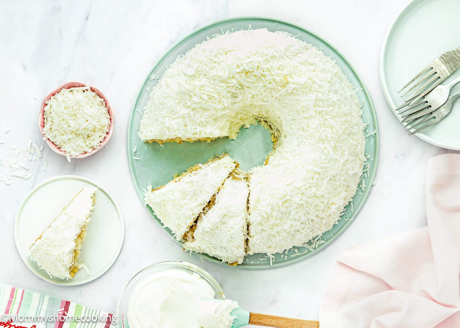 A round white frosted Eggless Coconut Bund Cake covered with shredded coconut, with one slice removed and placed on a plate; a bowl of extra coconut and utensils are nearby.