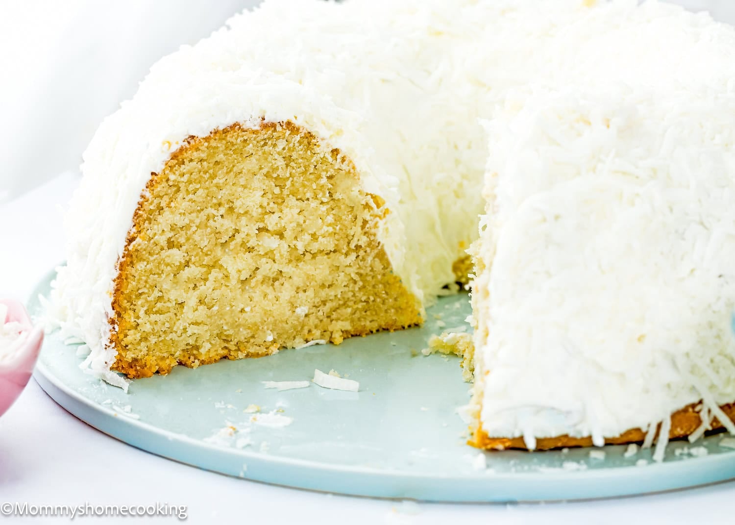A close-up of an Eggless Coconut Bundt Cake covered in white icing and shredded coconut, displayed on a light blue plate.