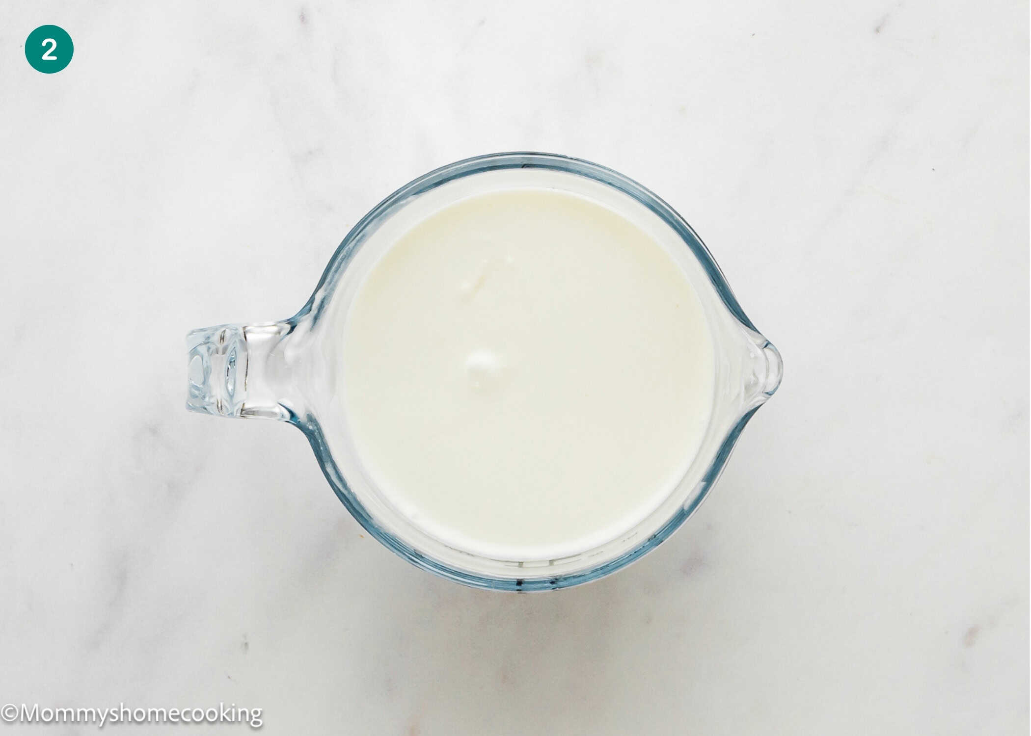 A glass measuring cup filled with a white liquid, perfect for making an Eggless Coconut Bundt Cake, is placed on a white marble surface.