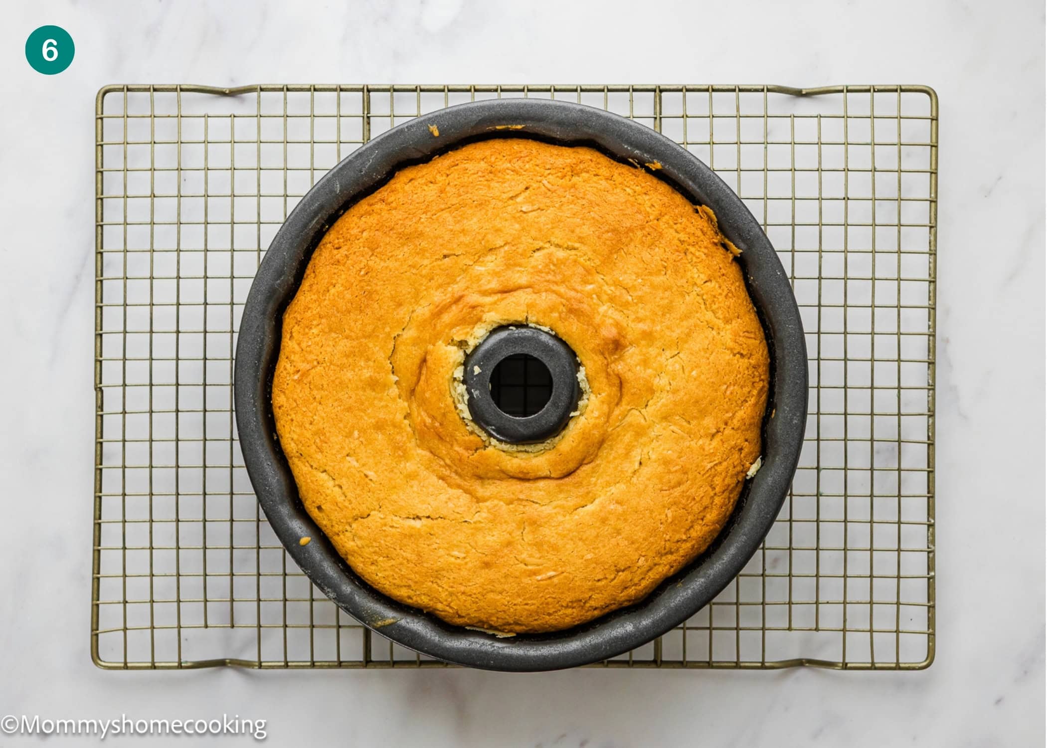 An Eggless Coconut Bundt Cake, golden-brown and freshly baked, sits in a bundt pan on a cooling rack over a white marble surface.