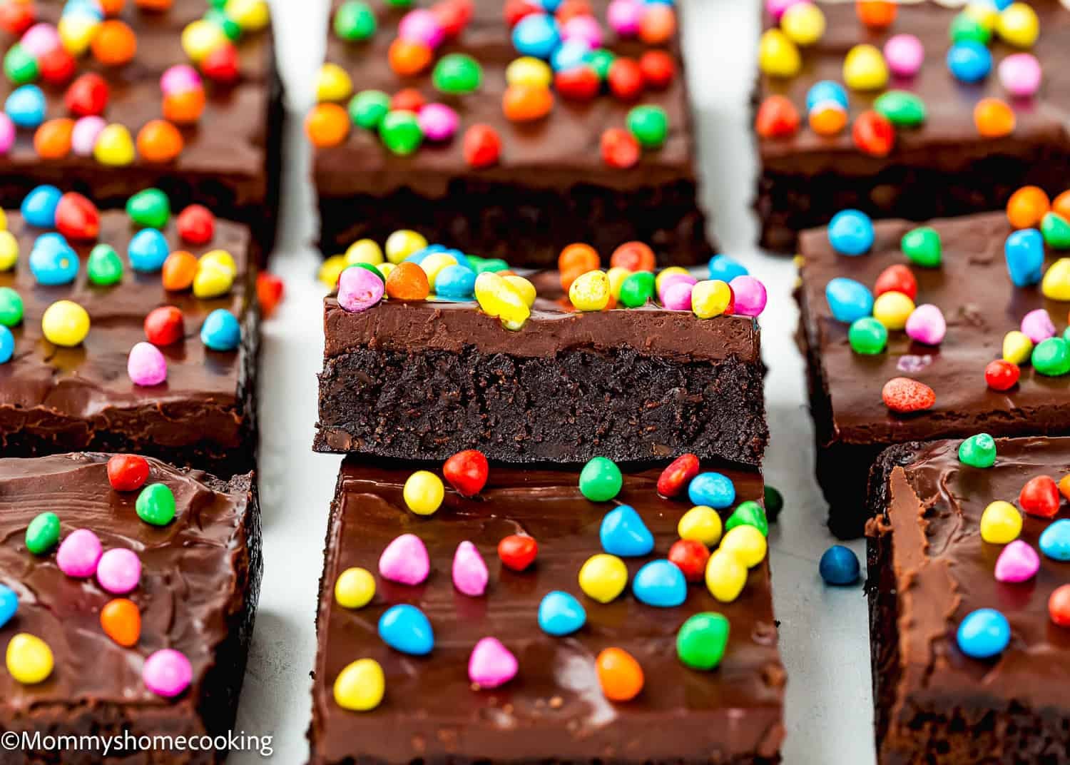 Eggless Cosmic Brownies topped with chocolate frosting and colorful candy pieces, arranged in a grid with one brownie stacked on top.
