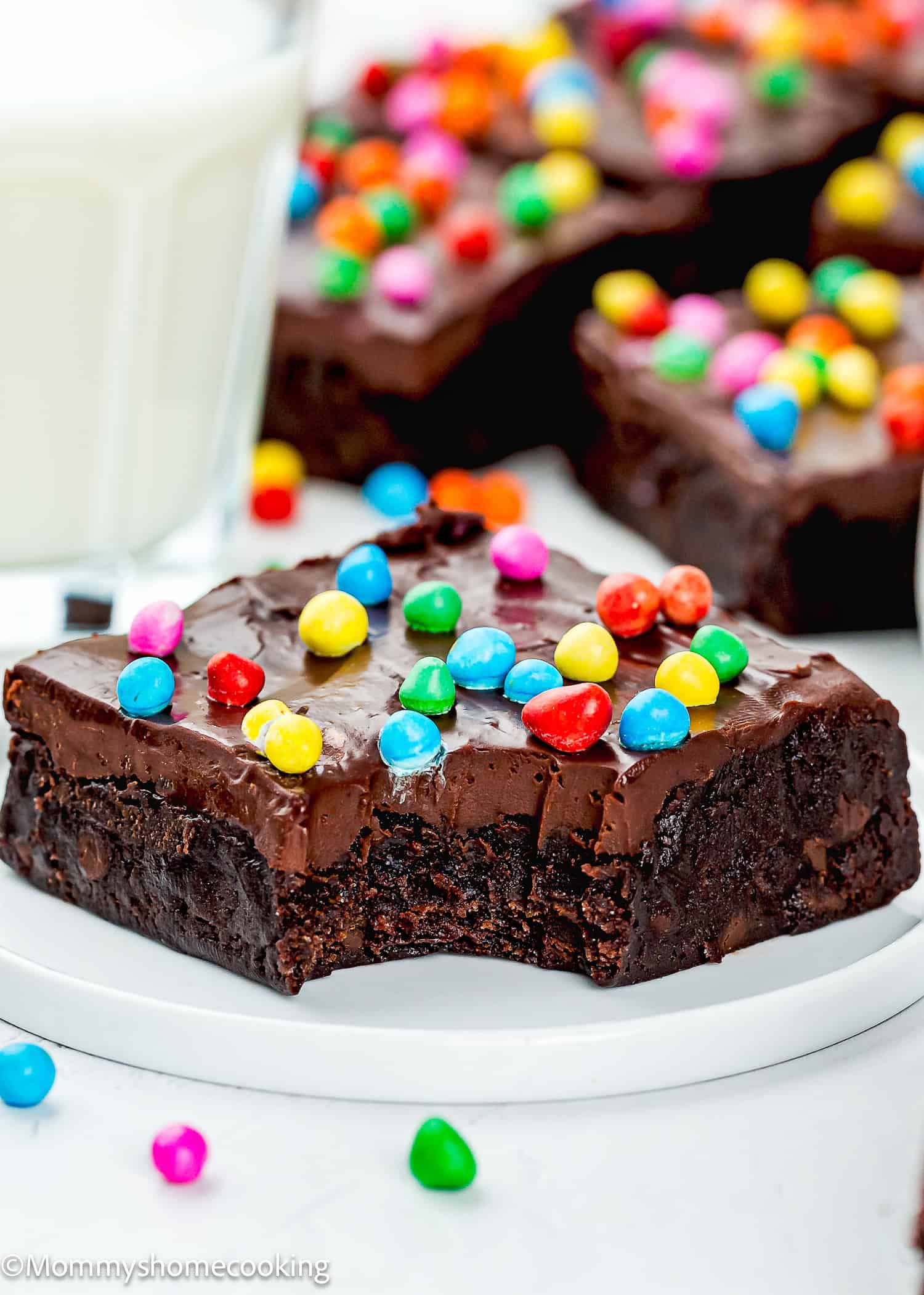 An Eggless Cosmic Brownie topped with colorful candy-coated chocolates sits on a white plate, with a bite taken out of it. A glass of milk and more brownies are in the background.