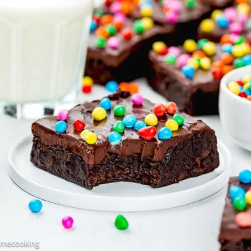 An Eggless Cosmic Brownie with a bite taken out, topped with colorful candy-coated chocolates, sits on a white plate beside a glass of milk and a small bowl of candies.