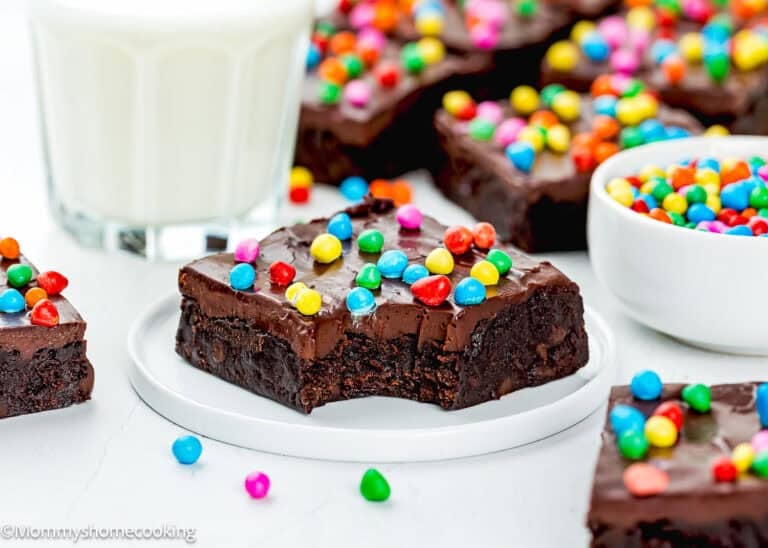 An Eggless Cosmic Brownie with a bite taken out, topped with colorful candy-coated chocolates, sits on a white plate beside a glass of milk and a small bowl of candies.