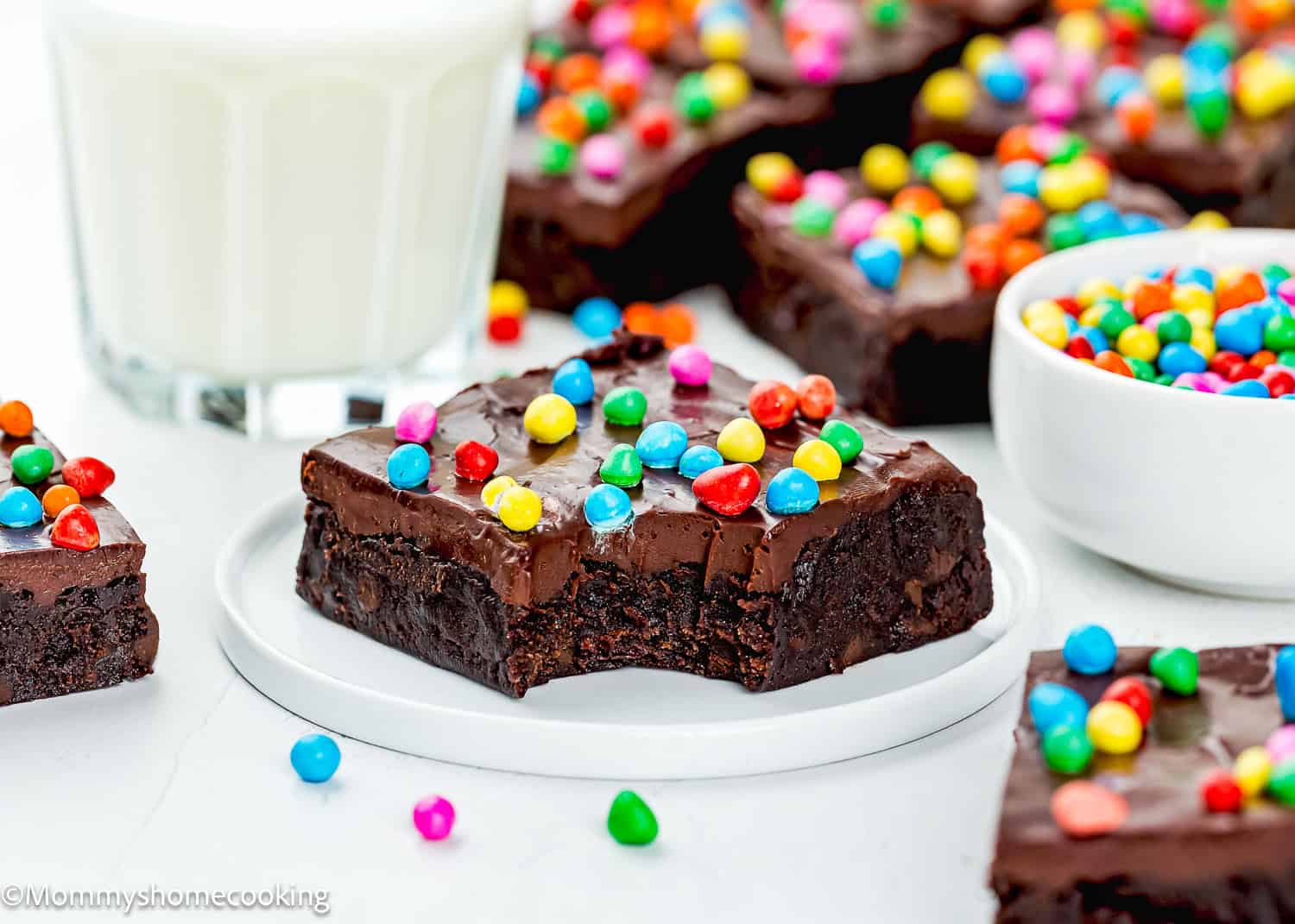 An Eggless Cosmic Brownie with a bite taken out, topped with colorful candy-coated chocolates, sits on a white plate beside a glass of milk and a small bowl of candies.