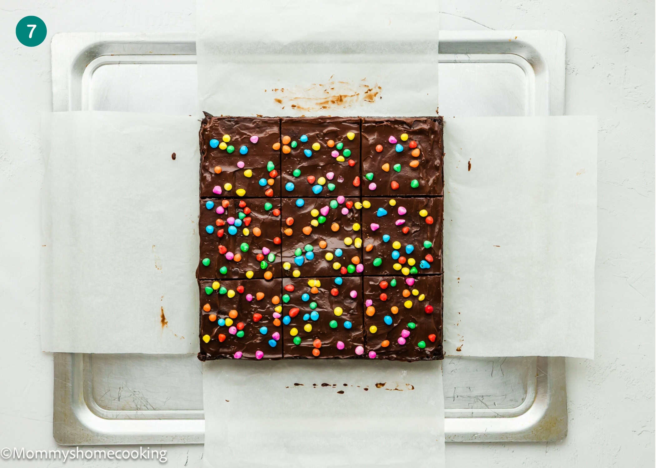 A tray of Eggless Cosmic Brownies topped with colorful candy sprinkles, cut into 16 squares on parchment paper over a metal baking sheet.