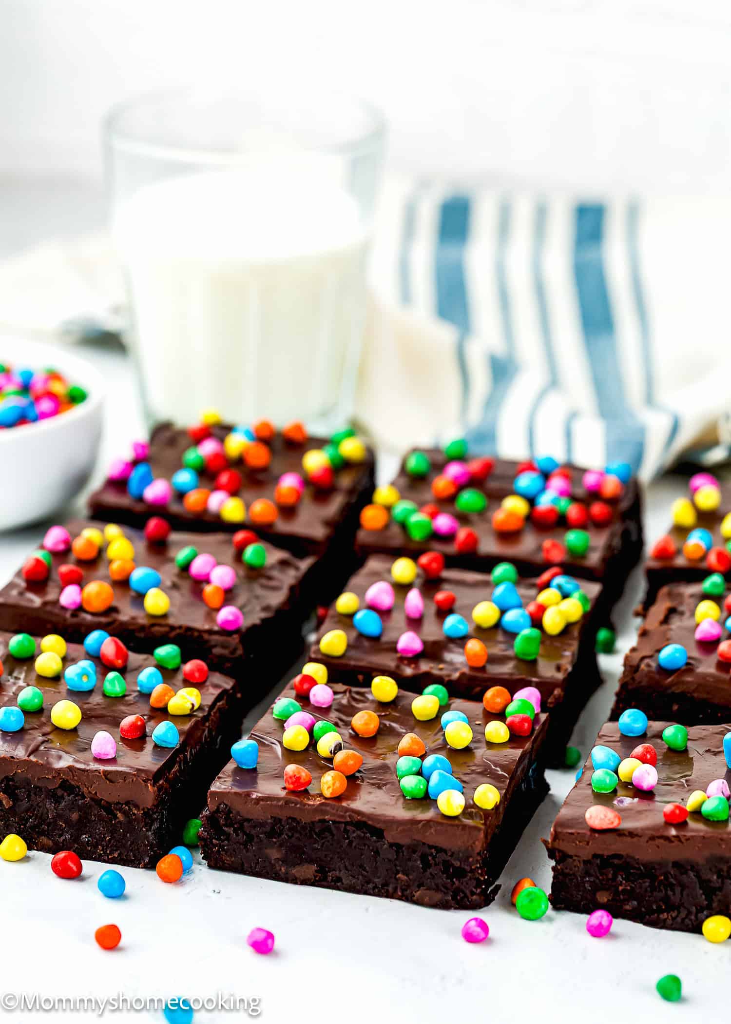 Eggless Cosmic Brownies topped with colorful candy pieces are arranged in rows next to a glass of milk and a striped cloth.