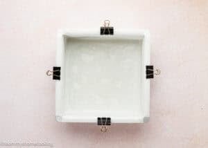 A square baking pan lined with parchment paper and secured at the edges with four binder clips, perfect for preparing an Eggless Hostess Cake, viewed from above on a light surface.