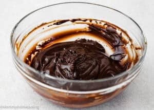 A glass bowl partially filled with thick, glossy melted chocolate on a light-colored surface, perfect for making Eggless Hostess Cake.
