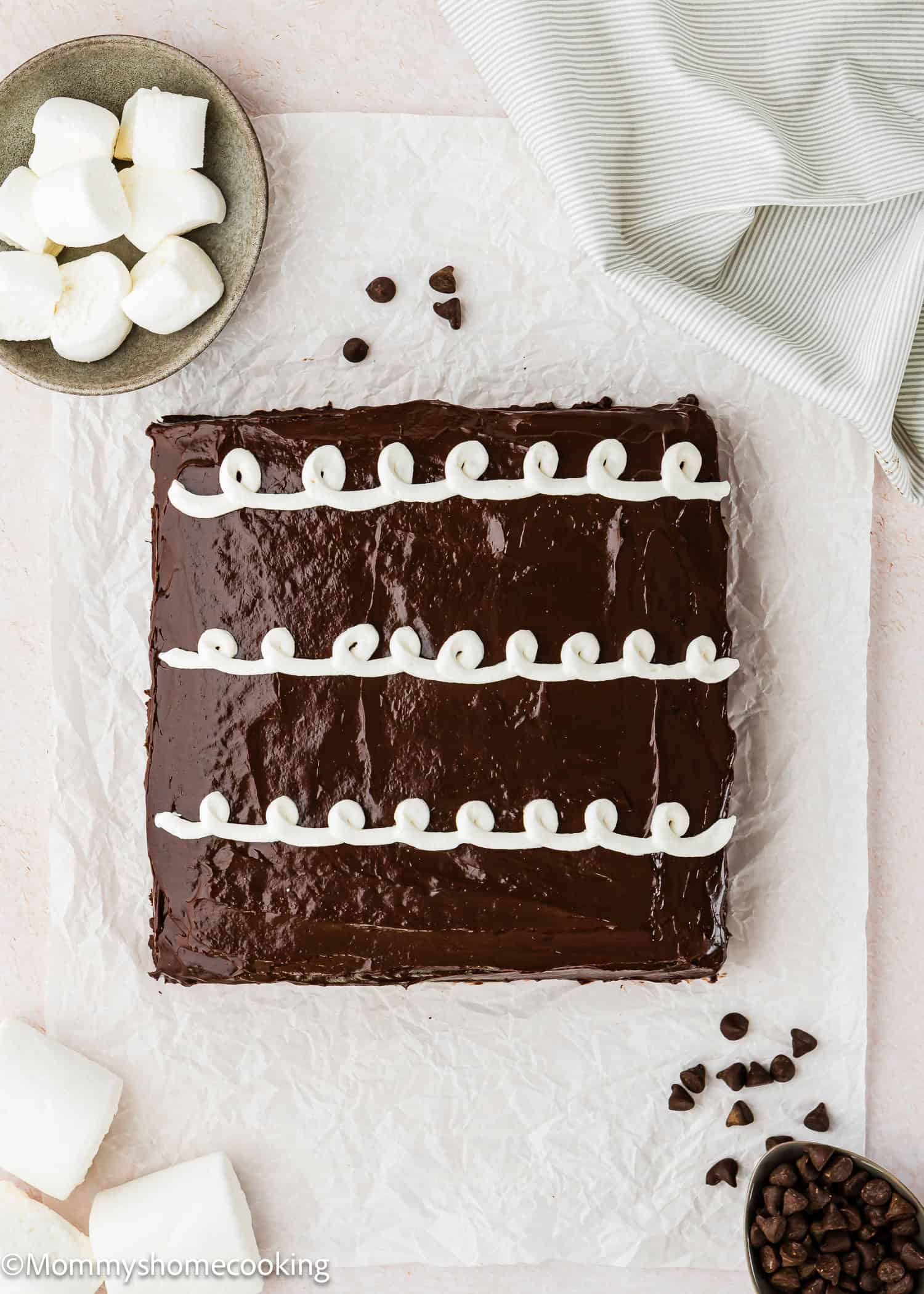 An Eggless Hostess Cake, square-shaped with three rows of white frosting swirls on top, sits on parchment paper surrounded by marshmallows and chocolate chips.