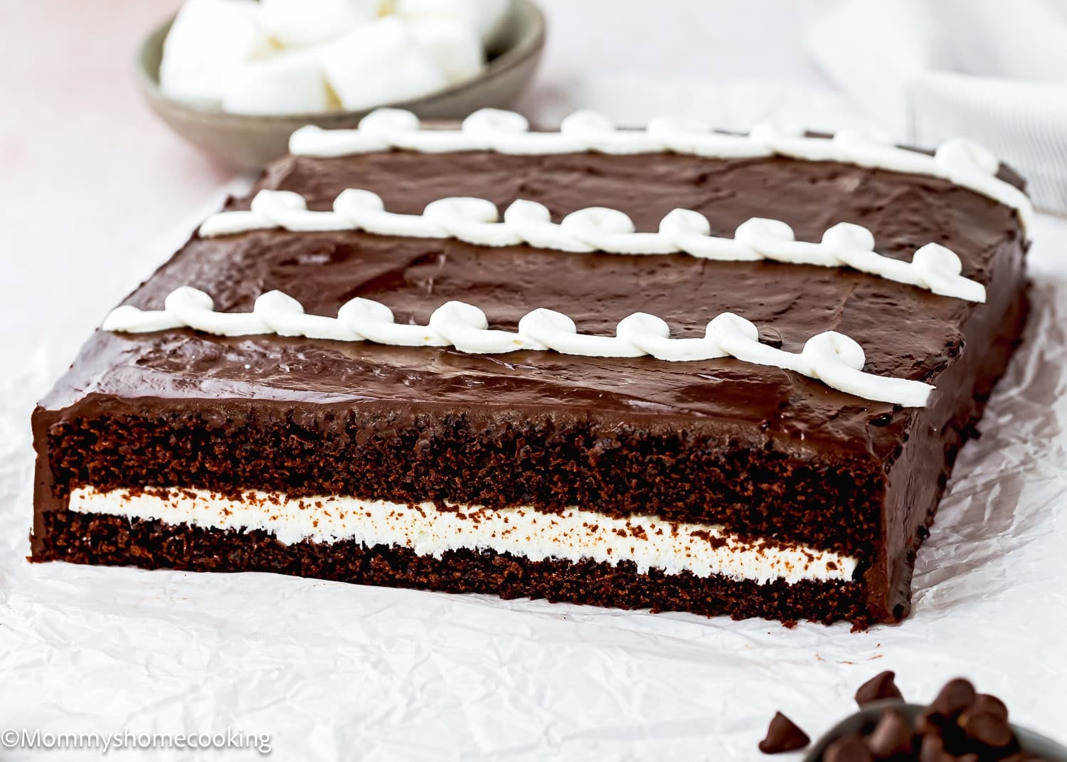 An Eggless Hostess Cake, rectangular with a white cream filling and chocolate frosting, decorated with white icing swirls on top, sits on parchment paper.