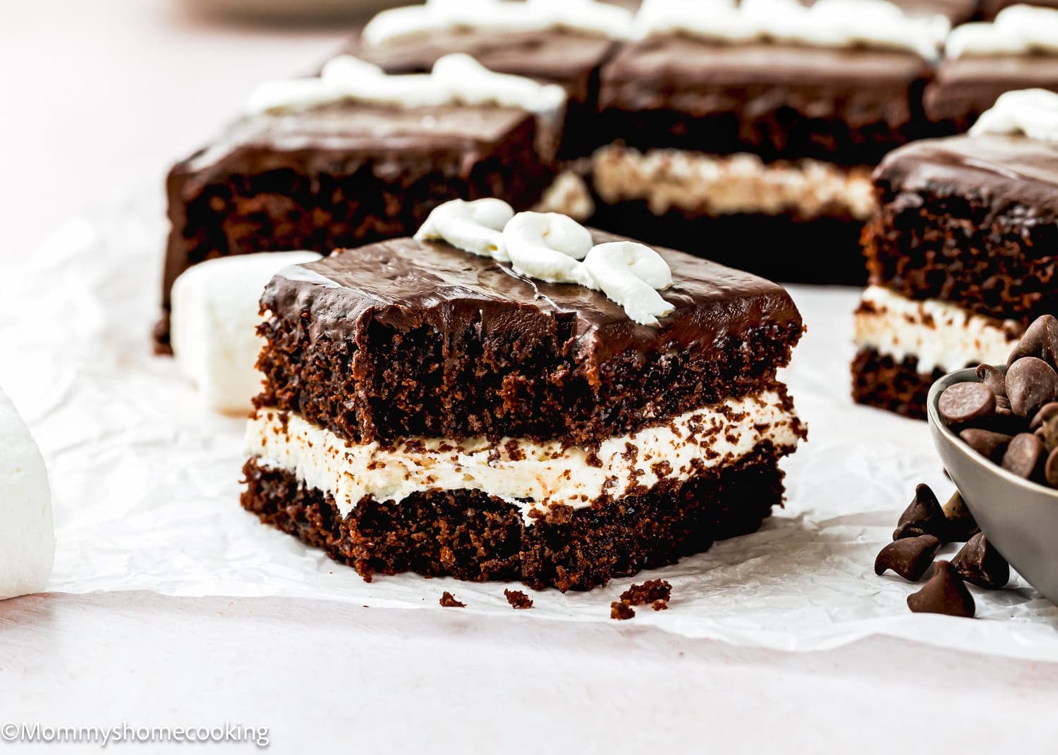 An Eggless Hostess Cake with a cream filling and chocolate frosting, topped with white icing, sits on parchment paper near chocolate chips and other cake pieces.