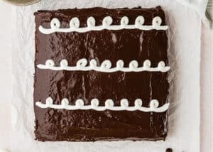 An Eggless Hostess Cake, this square chocolate cake features glossy frosting and three rows of white icing swirls on top, set on parchment paper.