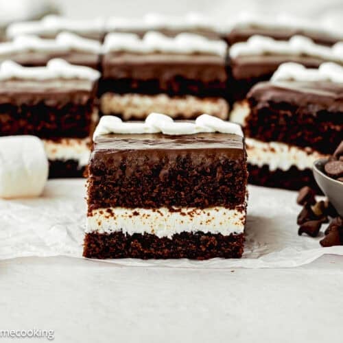 A close-up of an Eggless Hostess Cake with a white cream filling, topped with chocolate glaze and white frosting, surrounded by marshmallows and chocolate chips.