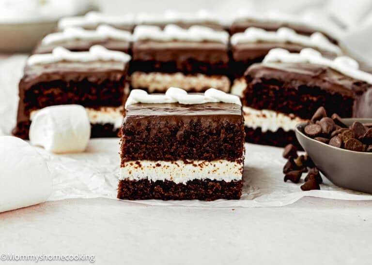 A close-up of an Eggless Hostess Cake with a white cream filling, topped with chocolate glaze and white frosting, surrounded by marshmallows and chocolate chips.