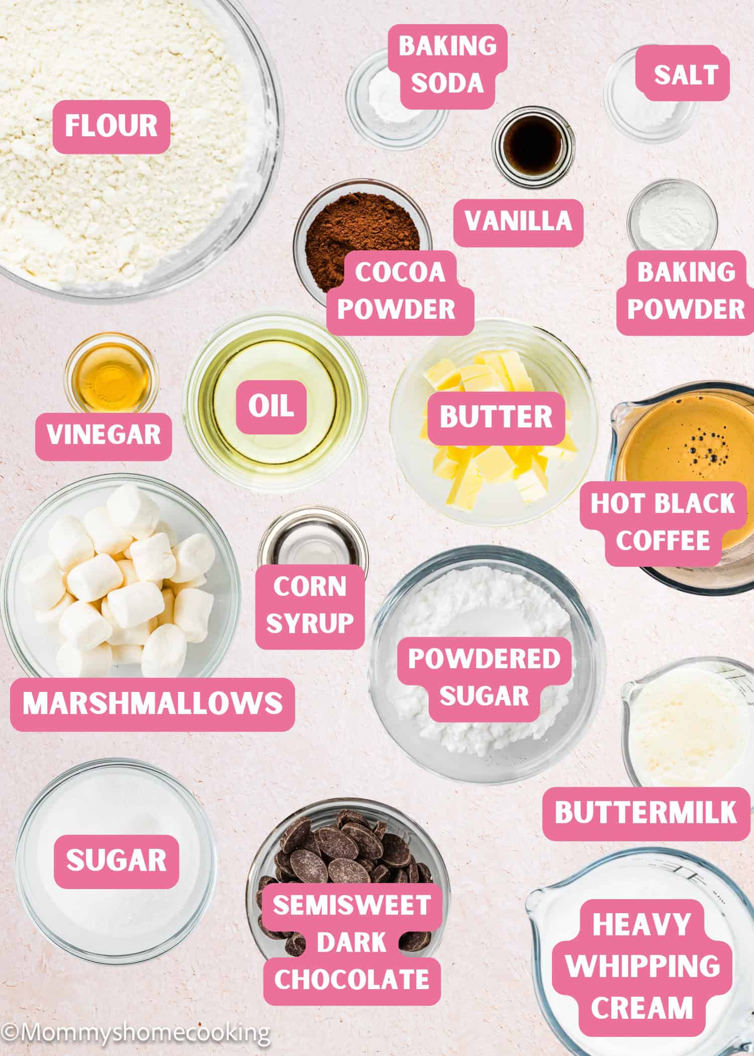 Various baking ingredients such as flour, baking soda, cocoa powder, vanilla, butter, marshmallows, chocolate, sugar, and oils are arranged in clear bowls on a light surface with labels—perfect for making an Eggless Hostess Cake.
