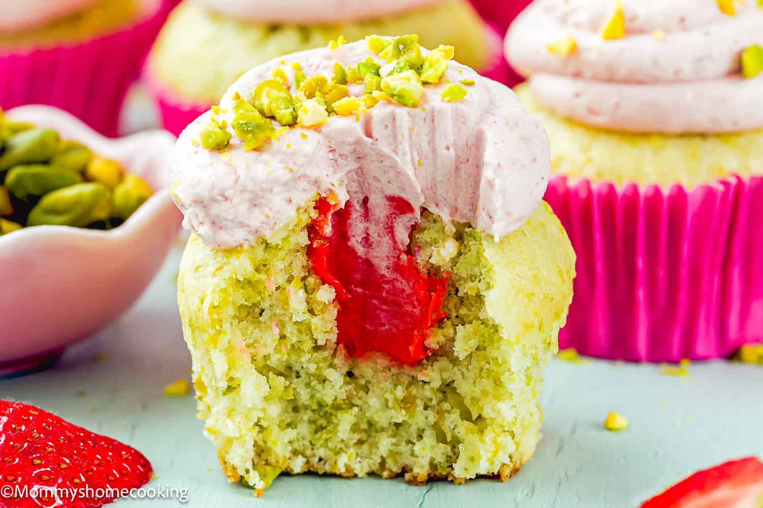 A partially eaten Eggless Pistachio Cupcake with pink strawberry buttercream and chopped pistachios on top, revealing a luscious white chocolate strawberry ganache filling inside.