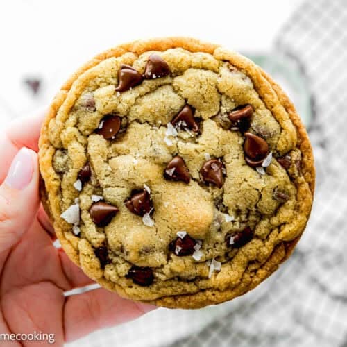 A hand holds a single serve chocolate chip cookie with visible chocolate chips and a sprinkle of sea salt on top. This no egg treat is in focus, while a checkered cloth is blurred in the background.
