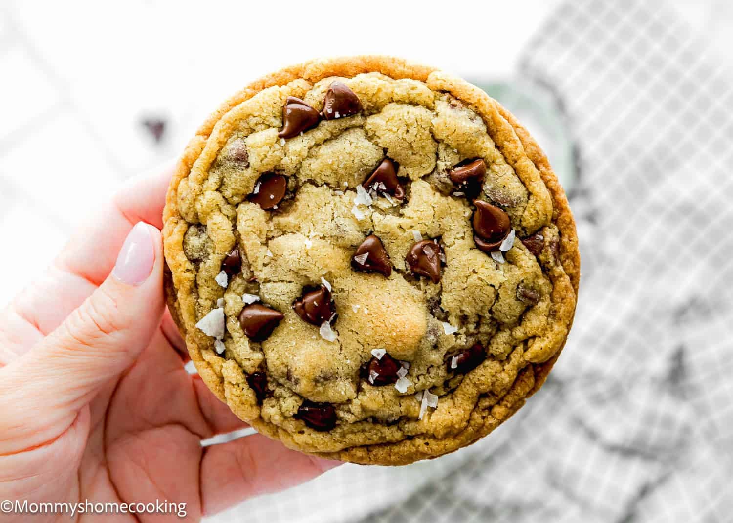 A hand holds a single serve chocolate chip cookie with visible chocolate chips and a sprinkle of sea salt on top. This no egg treat is in focus, while a checkered cloth is blurred in the background.