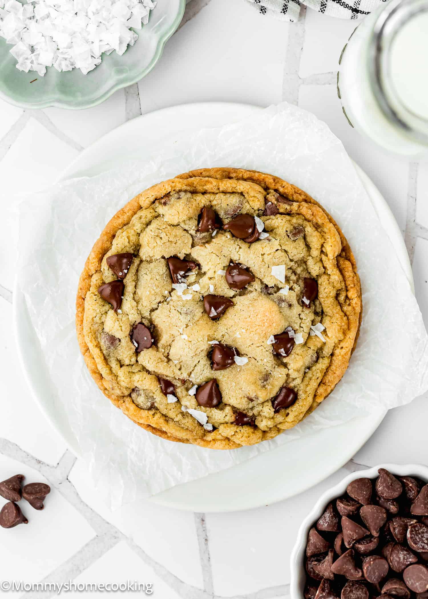 A single serve chocolate chip cookie no egg sits on parchment paper on a white plate, topped with sea salt flakes and surrounded by chocolate chips, coconut flakes, and a glass of milk.