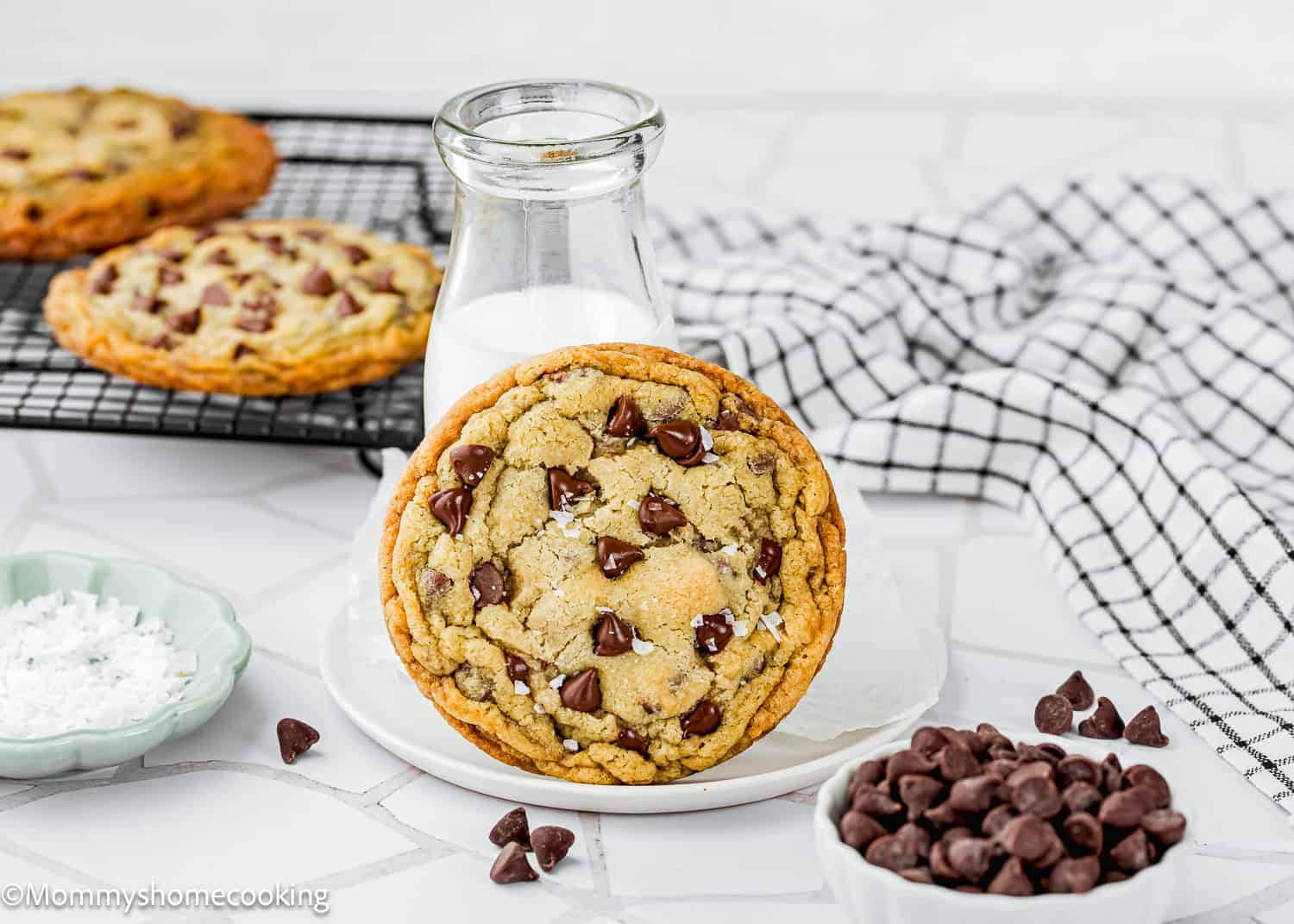 A Single Serve Chocolate Chip Cookie No Egg rests against a bottle of milk on a plate, with more cookies, chocolate chips, and a dish of salt nearby. A checkered cloth is in the background.