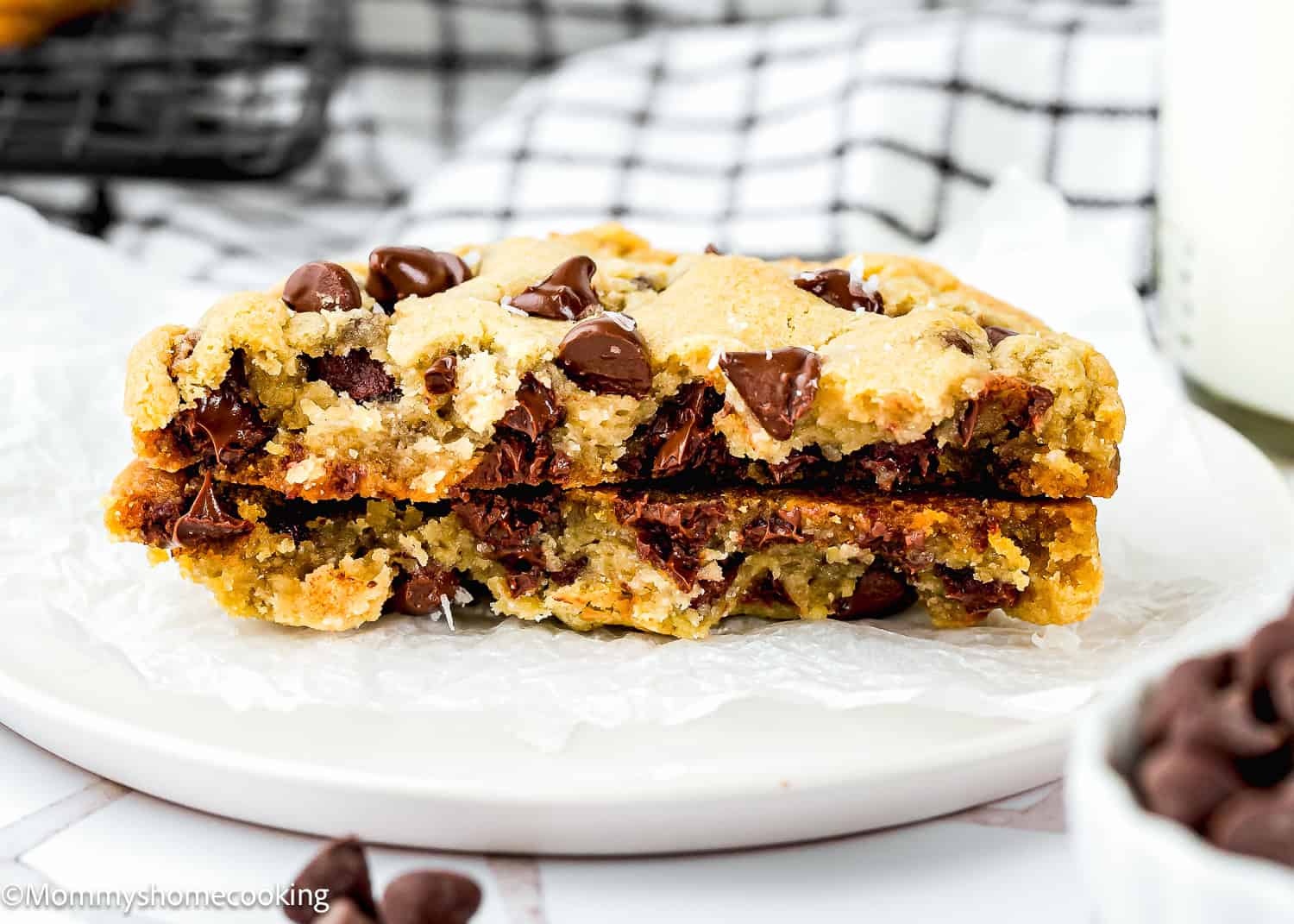 A thick Single Serve Chocolate Chip Cookie No Egg broken in half reveals melted chocolate chips inside, resting on a white plate next to a glass of milk and a checkered cloth in the background.