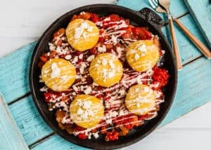 A skillet with six round yellow corn cakes on a bed of tomato and vegetable sauce, topped with white crumbly cheese and drizzled with a white sauce. Inspired by Venezuelan Bollos Pelones. Fork and knife on the side.