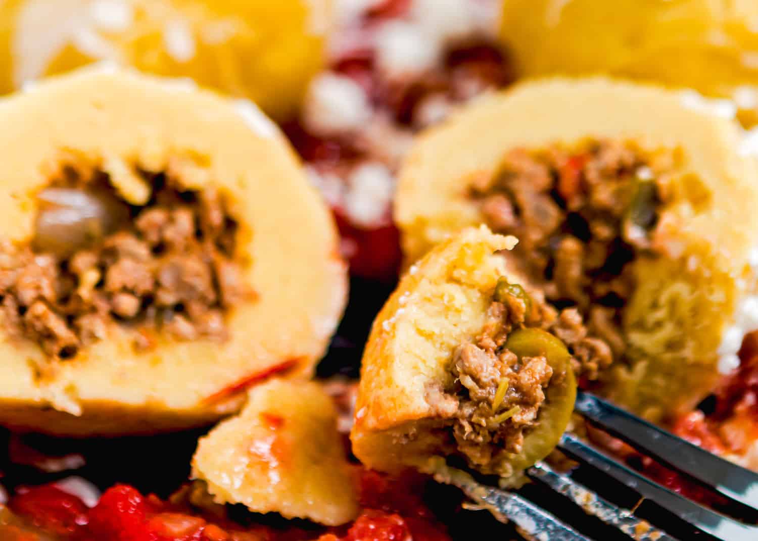 Close-up of a fork holding a piece of Venezuelan Bollos Pelones—stuffed potato ball filled with seasoned ground beef, surrounded by tomato sauce and more potato balls in the background.