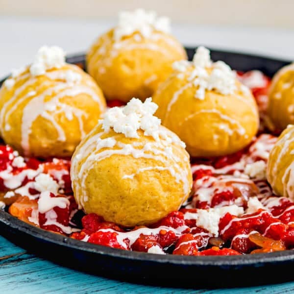 A black plate with six golden Venezuelan Bollos Pelones topped with white cheese and sauce, served over a bed of chopped tomatoes and peppers.