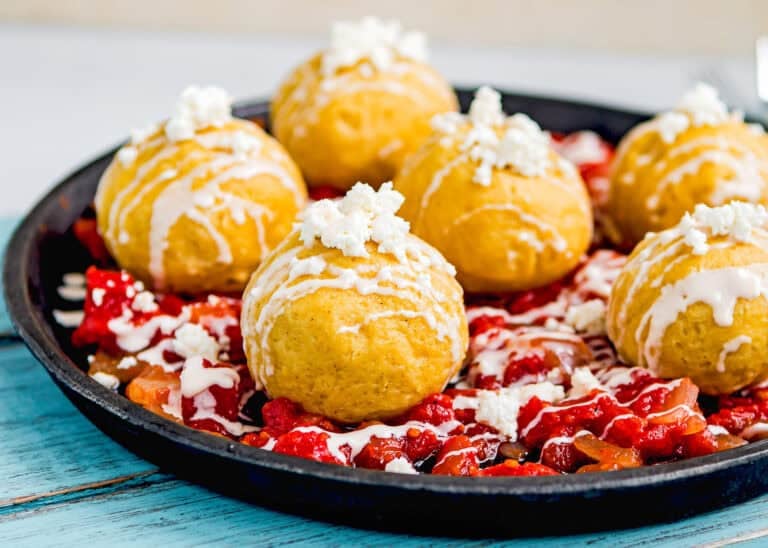 A black plate with six golden Venezuelan Bollos Pelones topped with white cheese and sauce, served over a bed of chopped tomatoes and peppers.