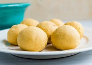 A white plate with several round, uncooked dough balls, known as Venezuelan Bollos Pelones. A turquoise bowl is partially visible in the background.
