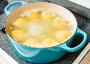 A blue pot filled with boiling water and Venezuelan Bollos Pelones dough balls cooking on a stovetop.