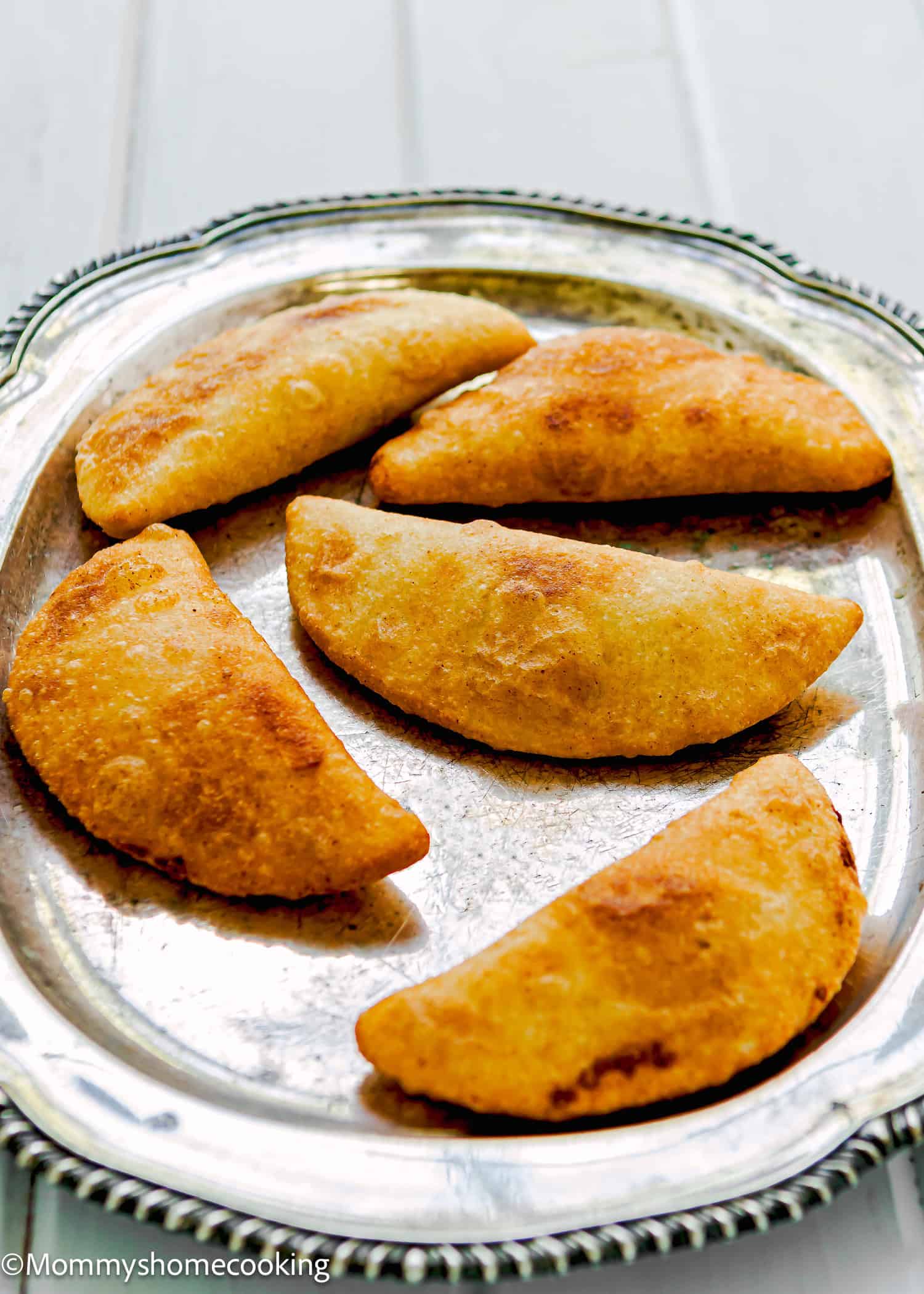 Five golden-brown Venezuelan Cazón Empanadas are arranged on a silver tray, displayed on a light-colored surface.