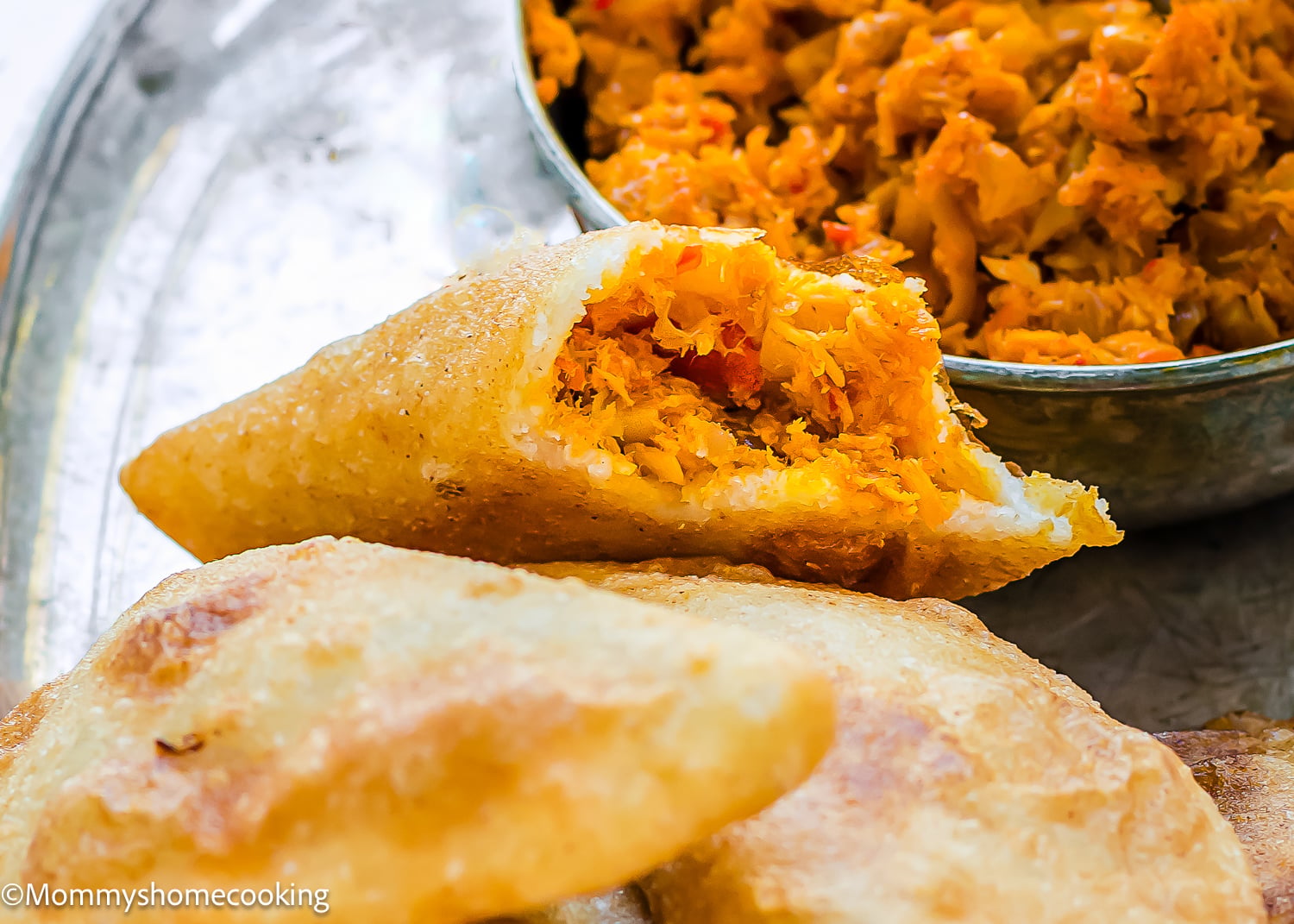 A close-up of Venezuelan Cazón Empanadas, fried and filled with a shredded vegetable mixture, with extra filling visible in a metal bowl in the background.