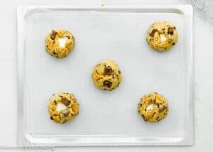 Five unbaked Egg-Free S’mores Cookies with chocolate chunks and marshmallows are arranged on a parchment-lined baking sheet.