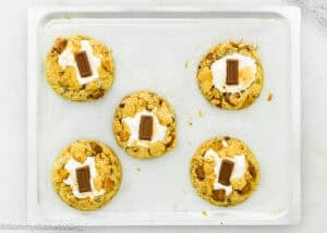 Five Egg-Free S’mores Cookies topped with marshmallows and chocolate pieces are arranged on a baking tray lined with parchment paper.