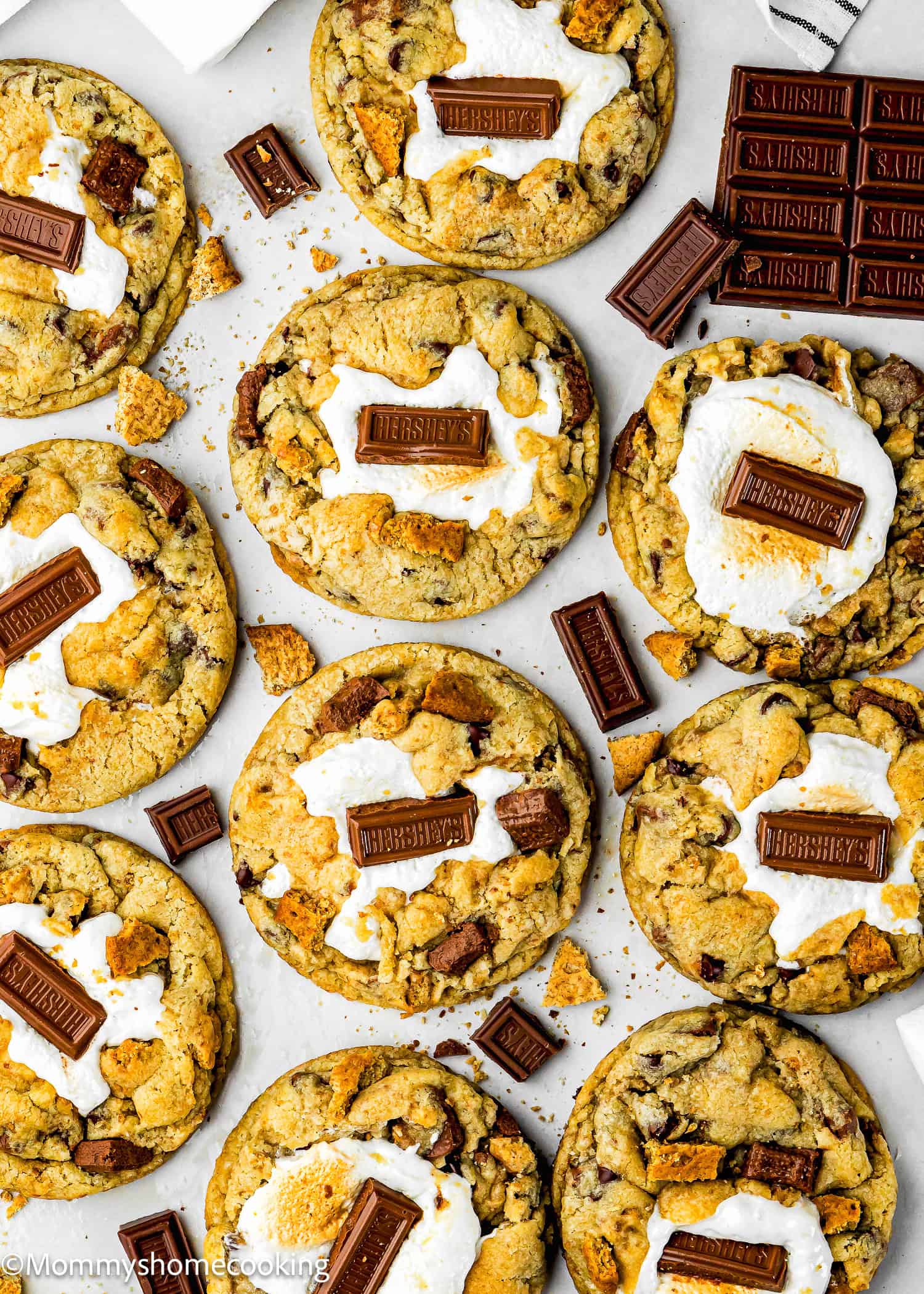 A flat lay of Egg-Free S’mores Cookies topped with toasted marshmallows, pieces of Hershey's chocolate, and crumbled graham crackers on a white surface.