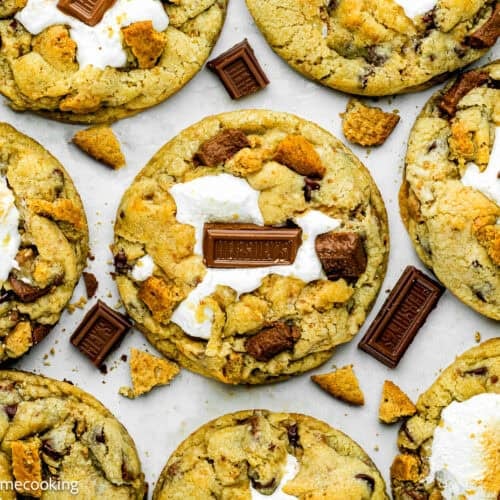 Several large Egg-Free S’mores Cookies topped with toasted marshmallows and pieces of milk chocolate sit on a white surface with some cookie crumbs around them.