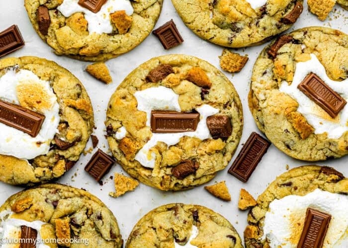 Several large Egg-Free S’mores Cookies topped with toasted marshmallows and pieces of milk chocolate sit on a white surface with some cookie crumbs around them.