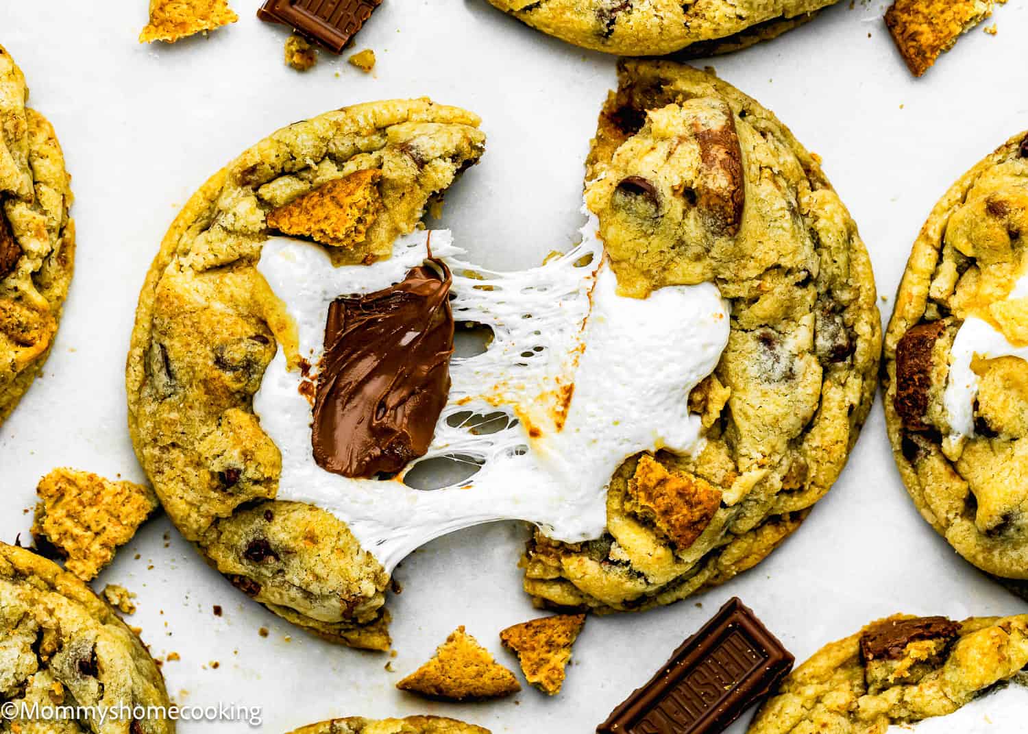 A close-up of gooey Egg-Free S’mores Cookies with melted chocolate and marshmallow filling, surrounded by cookie pieces and chocolate chunks on a white surface.