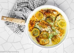 A round white pan with Eggless Chicken Francese fillets in a creamy sauce, garnished with lemon slices and chopped herbs, sits on a tiled surface next to a striped kitchen towel.