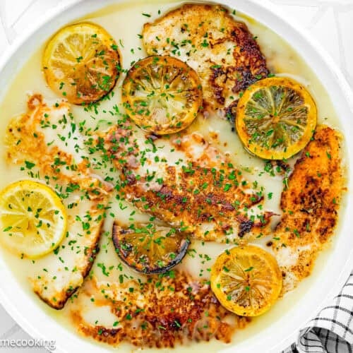 A skillet with browned Eggless Chicken Francese fillets in a light lemon sauce, garnished with parsley and sliced cooked lemons, set on a white tile surface next to a checkered cloth.