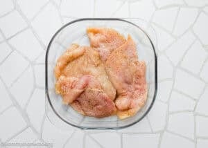 Raw, seasoned chicken breast fillets for Eggless Chicken Francese arranged in a glass baking dish on a white tiled surface.